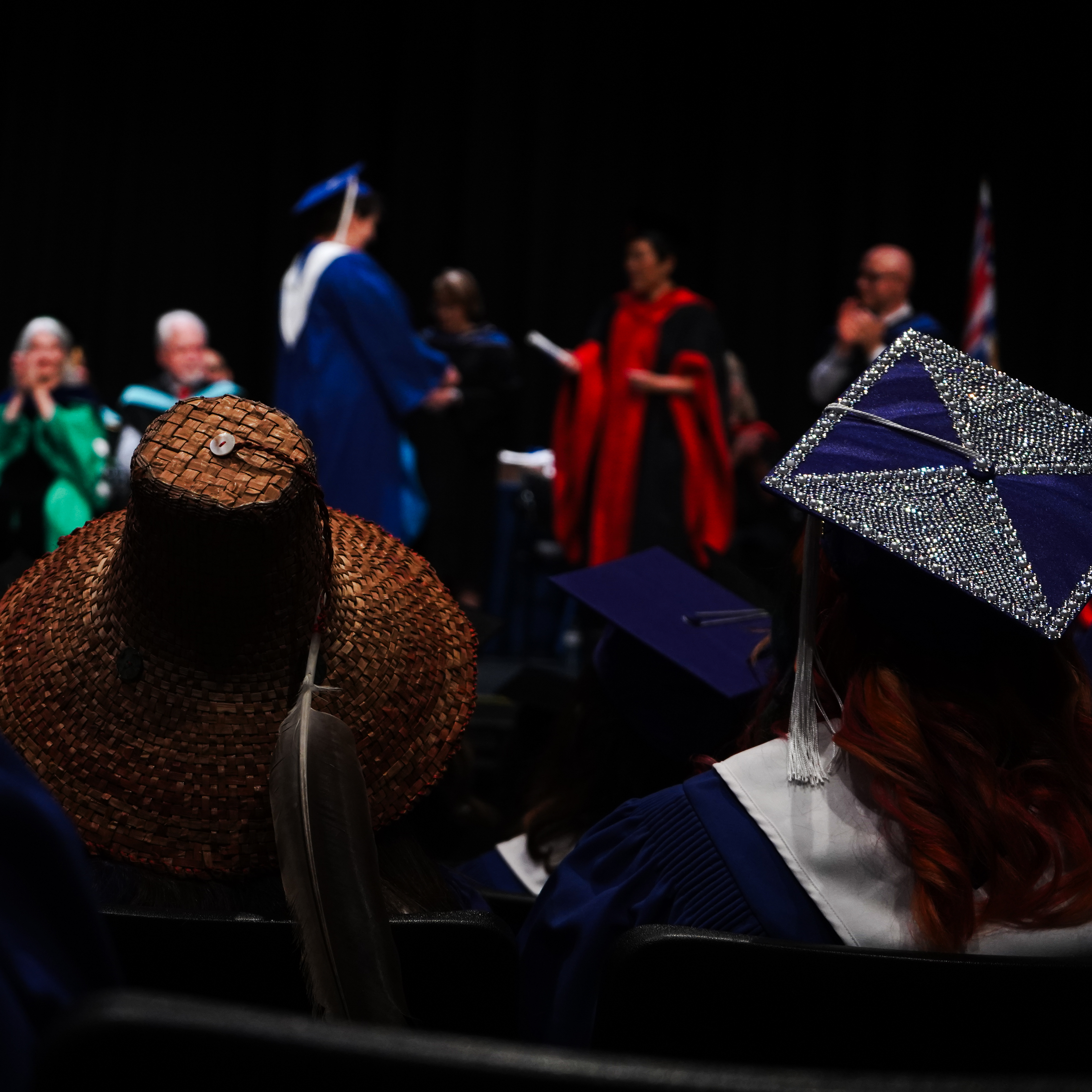 Two graduates in regalia watch from the audience as a fellow classmate crosses the stage.